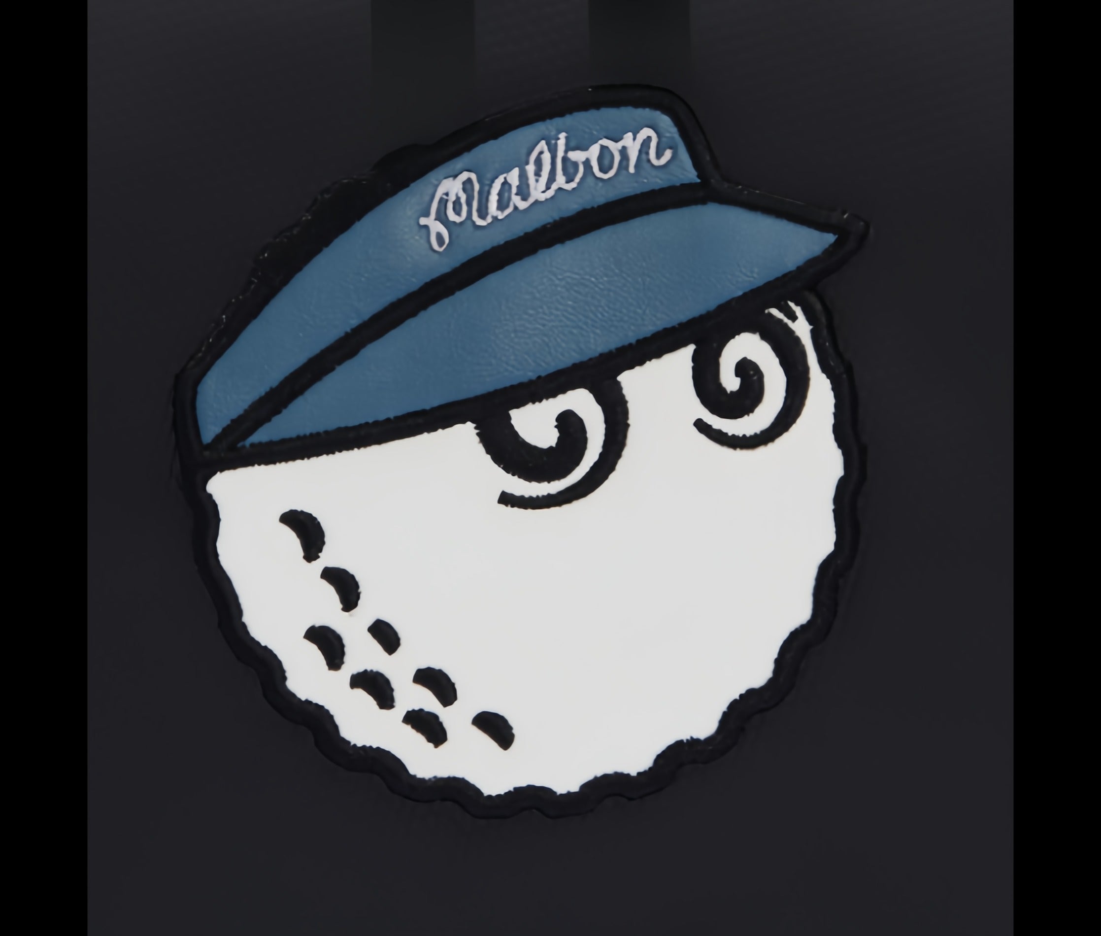 Detailed close-up of Malbon Golf mini handbag's embroidered mascot patch – white golf ball with cartoon eyes, navy blue cap with "Malbon" in white script, and black outline