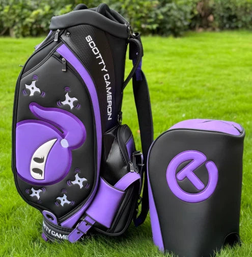 A full view of the Scotty Cameron Camo Collage Tour Bag in black and purple, featuring a unique ninja graphic and "Circle T" logo. The bag is displayed on a lush green golf course, showcasing its premium design and durable construction.